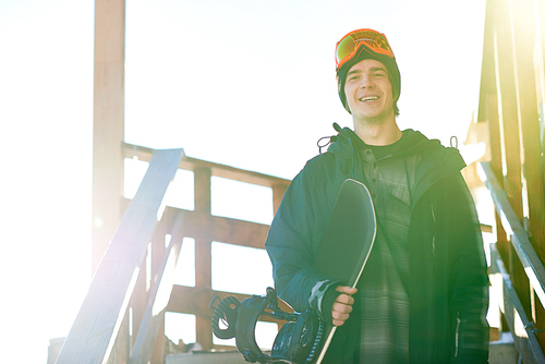 Portrait of modern young snowboarder smiling happily enjoying sunny winter day at ski resort exiting wooden chalet house, copy space