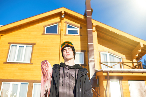 Low angle portrait of handsome young snowboarder  posing against wooden chalet at modern ski resort enjoying sunny winter, copy space