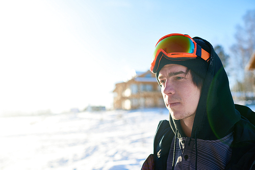 Portrait of handsome young snowboarder looking away enjoying sunny summer day at ski resort with wooden chalets in background, copy space