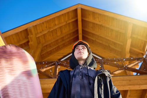 Low angle portrait of handsome young snowboarder posing against wooden chalet at modern ski resort enjoying sunny winter, copy space