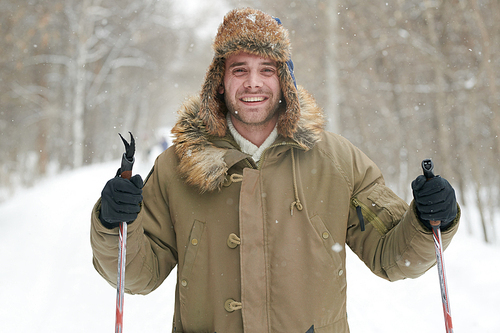 Waist up portrait of happy young man smiling at camera while enjoying skiing in snowy winter forest