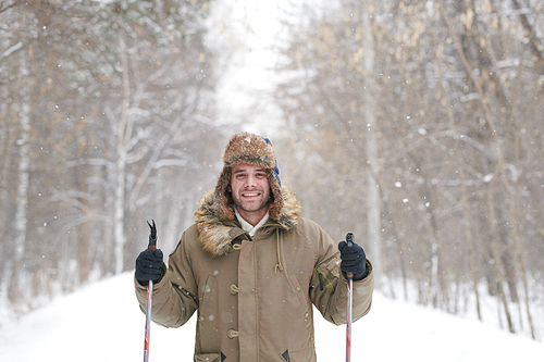 Waist up portrait of happy young man smiling at camera while enjoying skiing in snowy winter forest, copy space