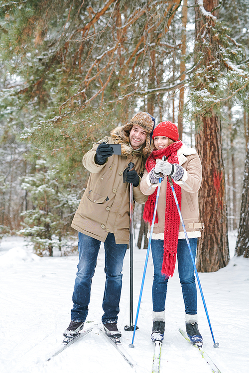 Full length portrait of active young couple taking selfie photo via smartphone while enjoying skiing in snowy winter forest during date