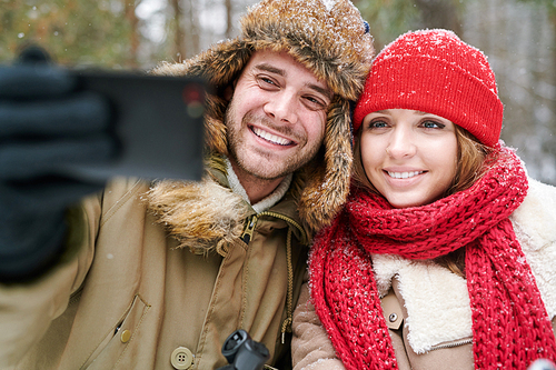 Portrait of happy young couple taking selfie photo via smartphone while enjoying date in winter forest