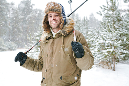 Waist up portrait of modern young man wearing fur hat smiling happily at camera while enjoying skiing in snowy winter park, copy space