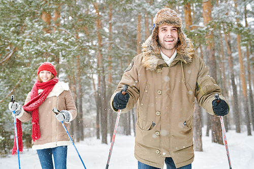 Waist up portrait of active young couple enjoying skiing in snowy winter forest, focus on smiling man in front, copy space