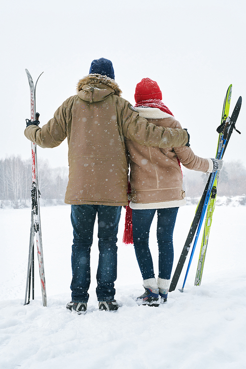 Full length back view portrait of loving young couple embracing while looking at view of beautiful winter landscape taking break during skiing trip