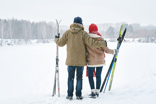 Full length back view portrait of loving young couple embracing while looking at view of beautiful winter landscape taking break during skiing trip, copy space