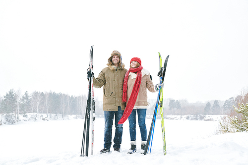 Full length portrait of loving young couple embracing and smiling happily while taking break during skiing trip and posing against beautiful winter landscape, copy space