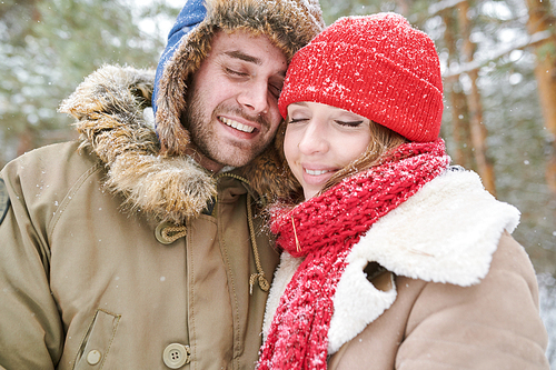 Head and shoulders portrait of loving young couple embracing tenderly and smiling with eyes closed while enjoying walk in snowy winter forest during date