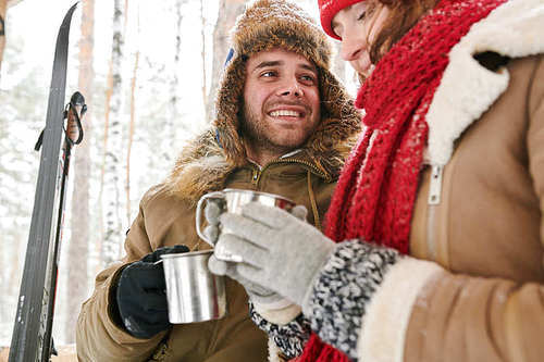 Waist up  portrait of loving young couple enjoying hot cocoa during date in beautiful winter forest, focus on smiling man looking at girlfriend, copy space