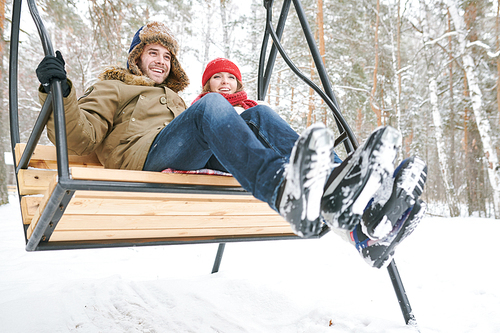 Full length low angle portrait of happy young couple sitting on wooden swing and laughing happily while enjoying date in winter forest, copy space