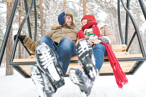 Full length low angle portrait of happy young couple sitting on wooden swing and looking at each other tenderly while enjoying date in winter forest