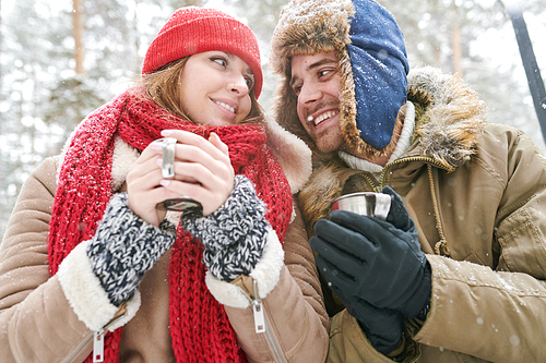 Low angle portrait of loving young couple enjoying hot cocoa during date in beautiful winter forest and looking at each other