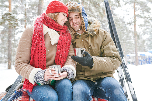 Portrait of loving young couple cuddling tenderly and  enjoying hot cocoa during date in beautiful winter forest, copy space