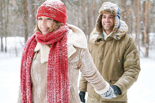 Waist up portrait of beautiful  young woman leading boyfriend through snowy winter forest during date, both smiling happily