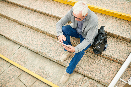 From above shot of senior man sitting on street steps near bag and using modern tablet