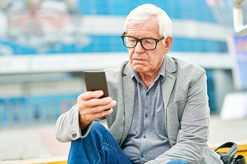 Senior man in glasses browsing modern smartphone while sitting on blurred background of street
