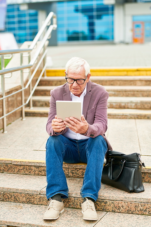 Elderly man in lilac jacket using modern tablet while sitting on street stairs near bag
