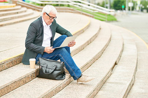 Side view of elderly man in glasses browsing modern laptop while sitting on street steps near bag and cup of hot beverage