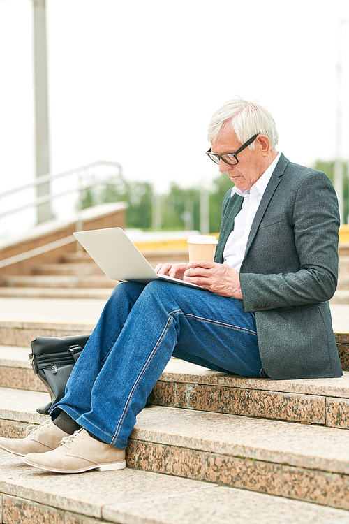 Side view of senior man holding cup of hot drink and browsing modern laptop while sitting on street stairs