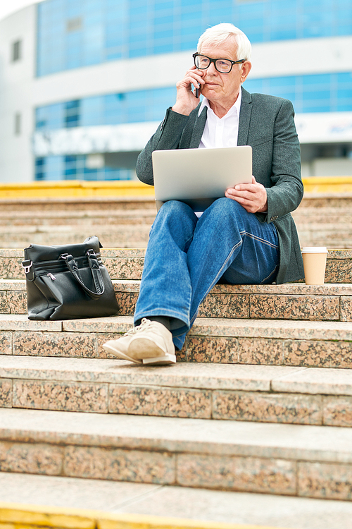 Aged man with modern laptop talking on smartphone while sitting on street stairs near bag and cup of hot beverage