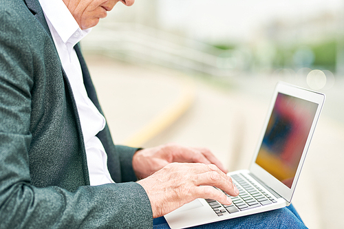 Side view of anonymous elderly man browsing modern laptop while sitting on blurred background of city street