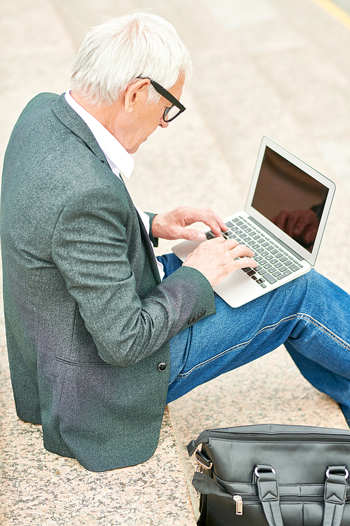 From above shot of elderly man in gray jacket browsing laptop while sitting on street stairs near bag