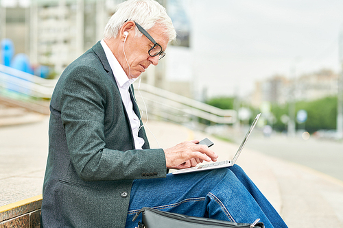 Side view of senior man listening to music in earphones and browsing modern laptop while sitting on blurred background of city street