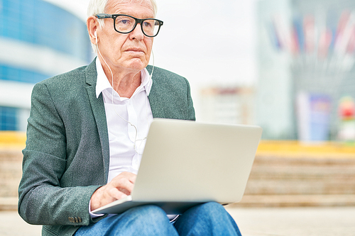 Aged man in glasses listening to music in earphones and browsing laptop while sitting on blurred background of street