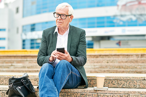Aged man in glasses browsing modern smartphone and listening to good music while sitting on street stairs near bag and cup of hot drink