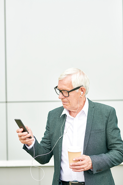 Aged man with cup of hot drink using smartphone and listening to music while standing near building on city street