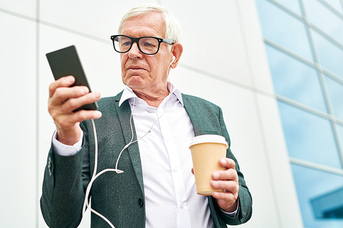 From below shot of aged man with cup of hot beverage browsing smartphone and listening to music while standing near modern building