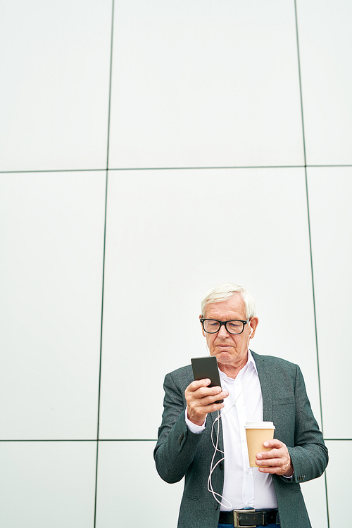 Senior man in glasses holding cup of hot beverage and listening to music while standing near wall of modern building and browsing smartphone