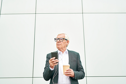 Elderly man in glasses holding cup of hot drink and looking away while using smartphone and listening to music on background of modern building wall