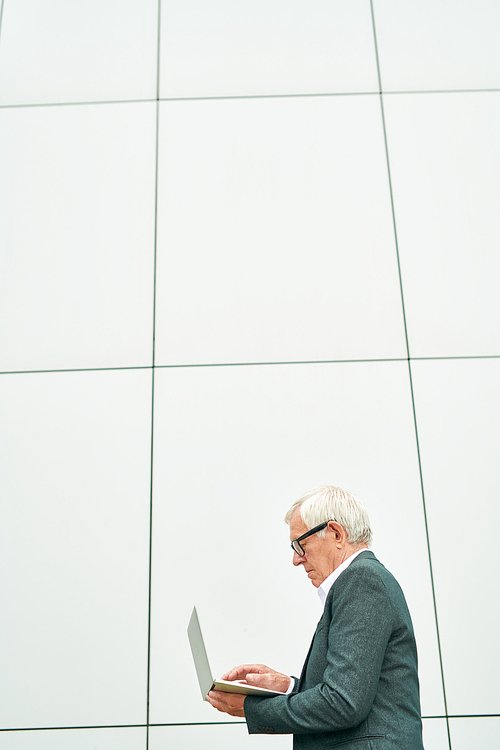 Side view of senior man standing near building of modern building and browsing laptop