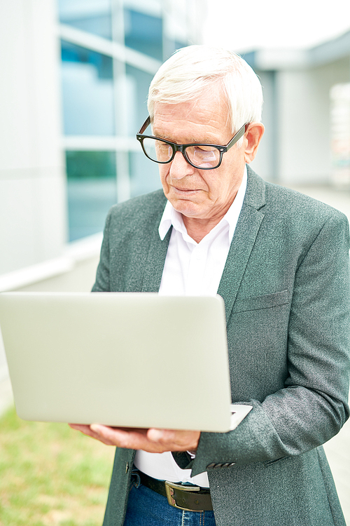 Senior man in glasses using modern laptop while standing on blurred background of building