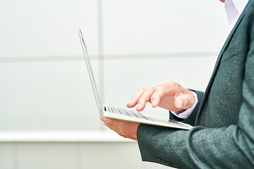 Side view of unrecognizable elderly man browsing laptop while standing near modern building