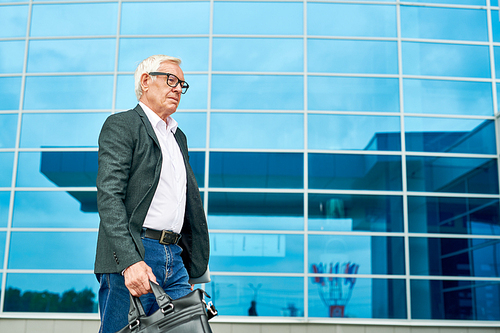 Side view of aged man with bag walking to work on background of modern office building
