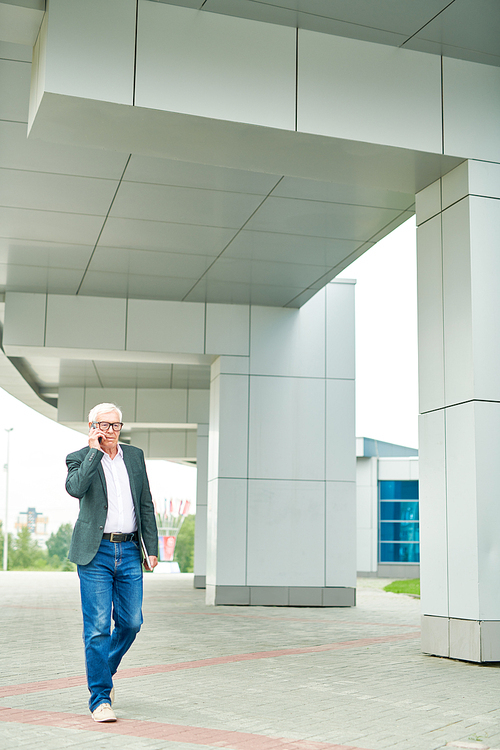Senior man having smartphone conversation while walking to work near modern building