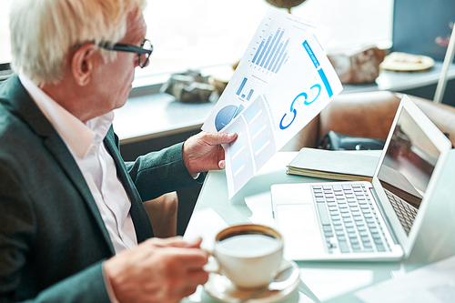 Side view of elderly man holding cup of aromatic coffee and examining documents with charts while sitting at cafe table near laptop