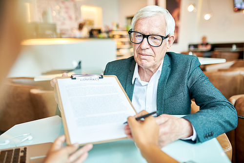 Portrait of successful senior businessman holding contract for signing during meeting in cafe