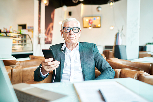 Portrait o modern senior businessman relaxing at table in cafe and listening to music from smartphone, copy space