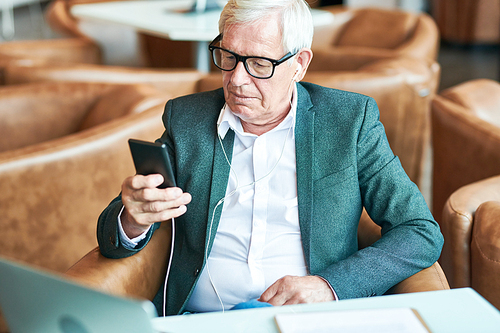 Portrait of modern senior businessman using smartphone while listening to music and  relaxing at table in cafe, copy space