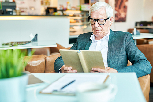 Portrait of successful senior businessman reading book in cafe while relaxing during coffee break, copy space, table