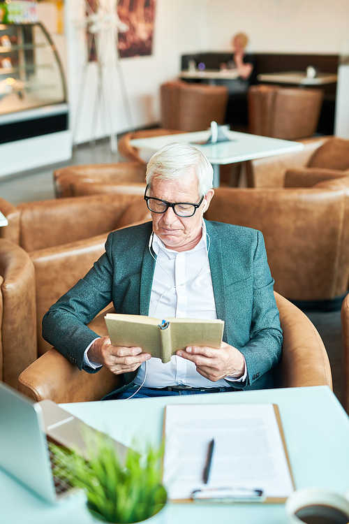 High angle portrait of successful senior businessman reading book in cafe while working during coffee break