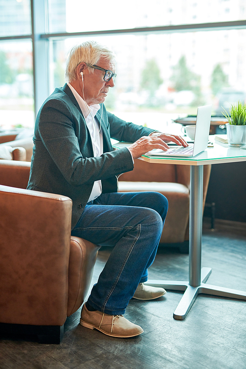 Side view full length portrait of senior man typing on keyboard while using laptop for browsing internet in cafe, copy space