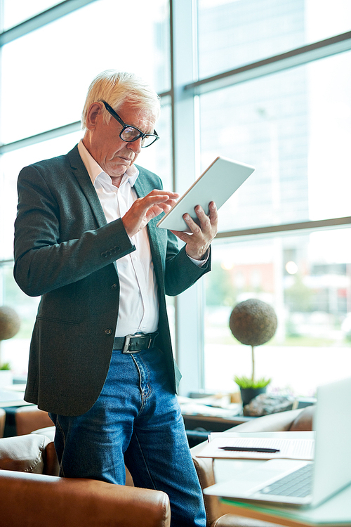 Side view portrait of successful senior businessman using digital tablet and browsing internet while standing at table in modern cafe