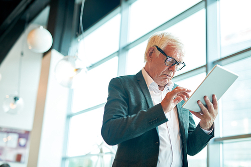 Low angle portrait of contemporary senior man using digital tablet and browsing internet while standing against window, copy space