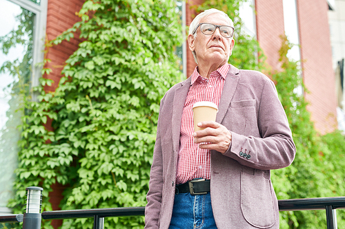 Low angle portrait of modern senior man dressed in business casual posing standing on terrace outdoors and holding paper coffee cup, copy space